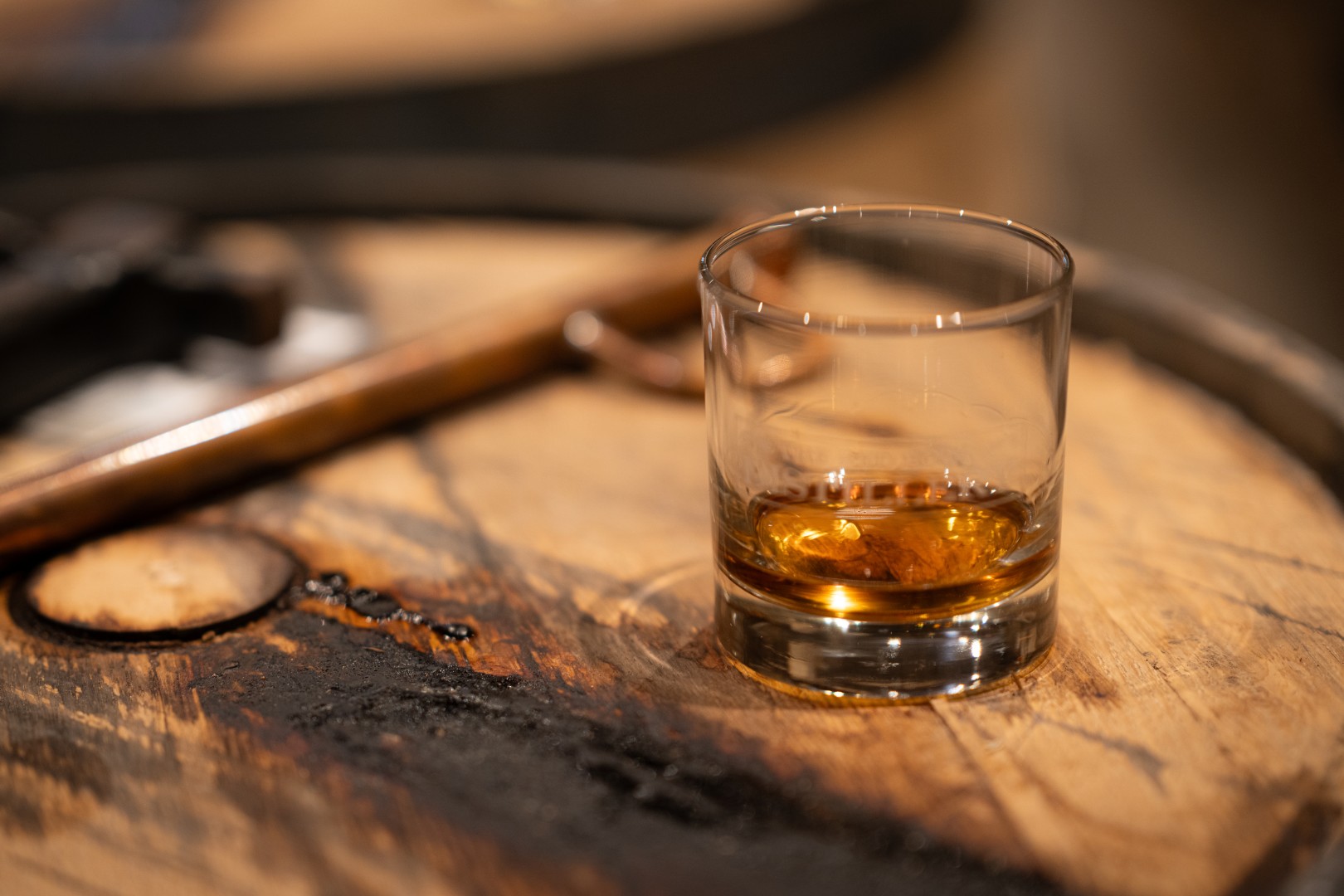A rocks glass with a small pour resting on a charred barrel head beside a copper valinch.