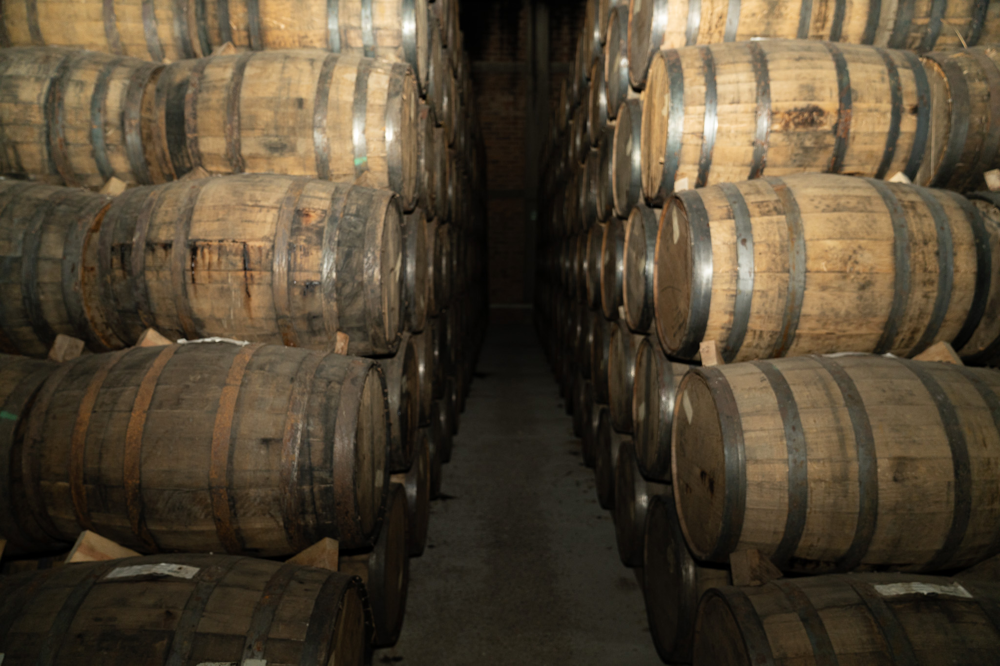 A corridor between stacked oak barrels in a dim rickhouse, warm light across the staves.